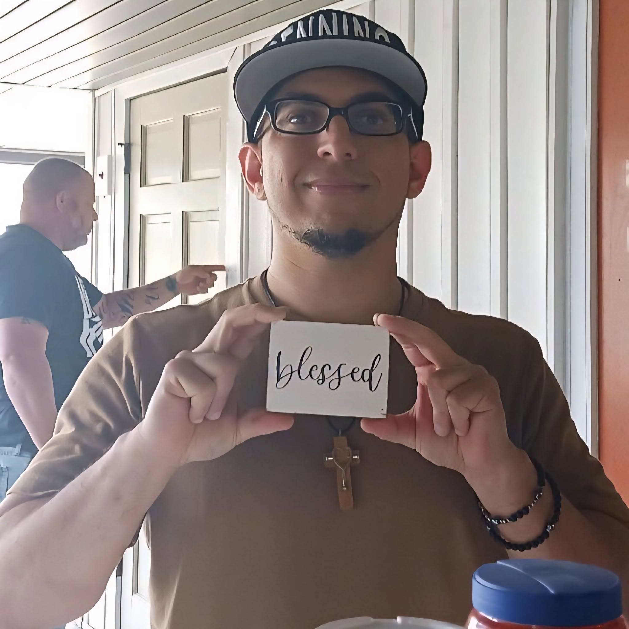Person holding a card with the word 'blessed' written on it, smiling, wearing glasses and a cap, indoors.