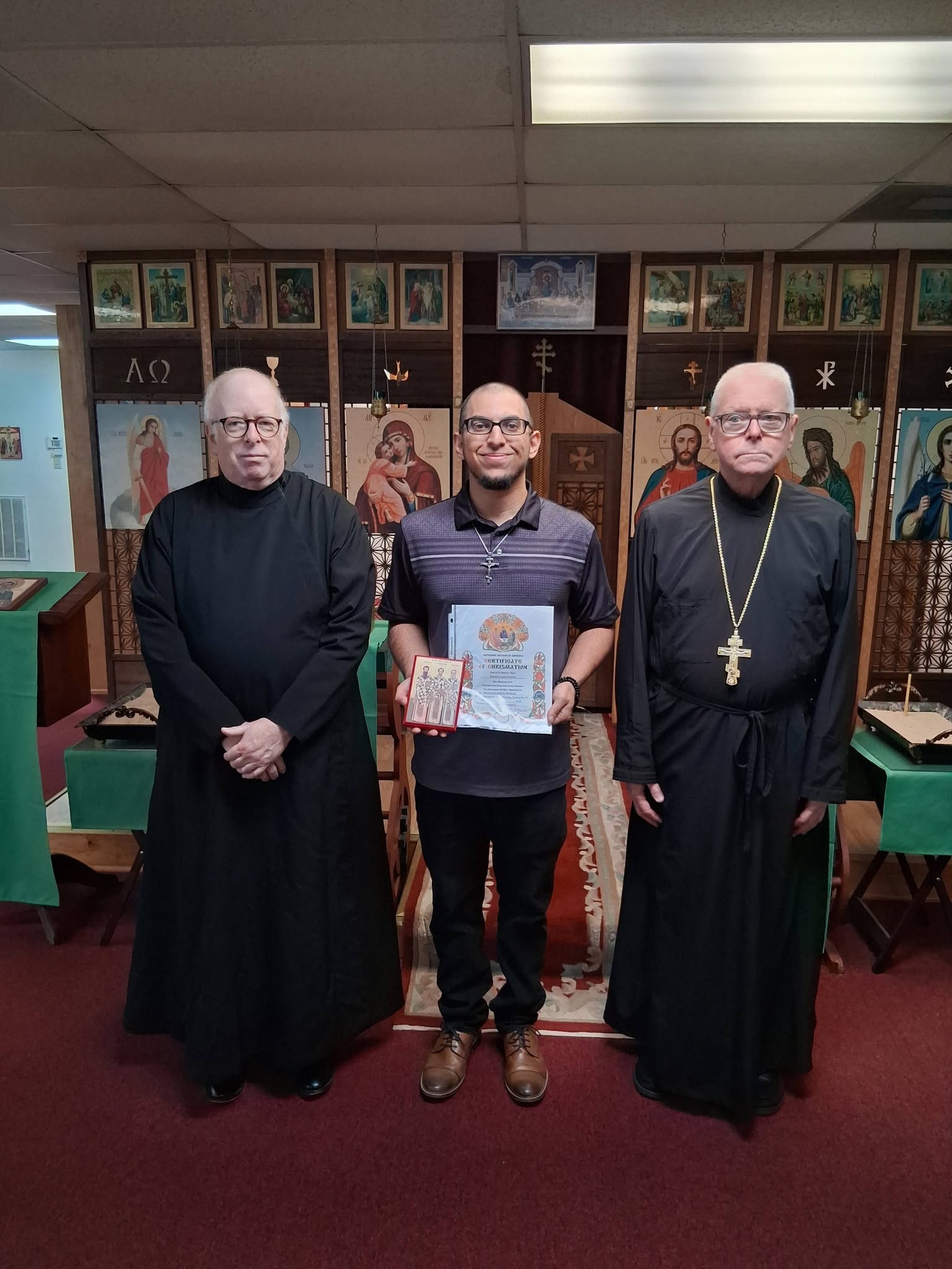 Three men standing in a room with religious icons and artwork on the wall, one holding a book or certificate.