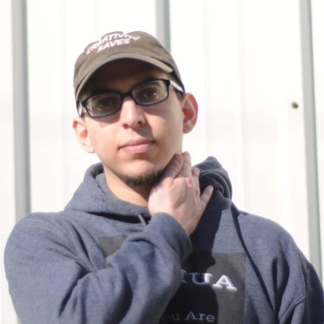 Young man wearing glasses, a cap, and a hoodie, standing outdoors in front of a white fence.