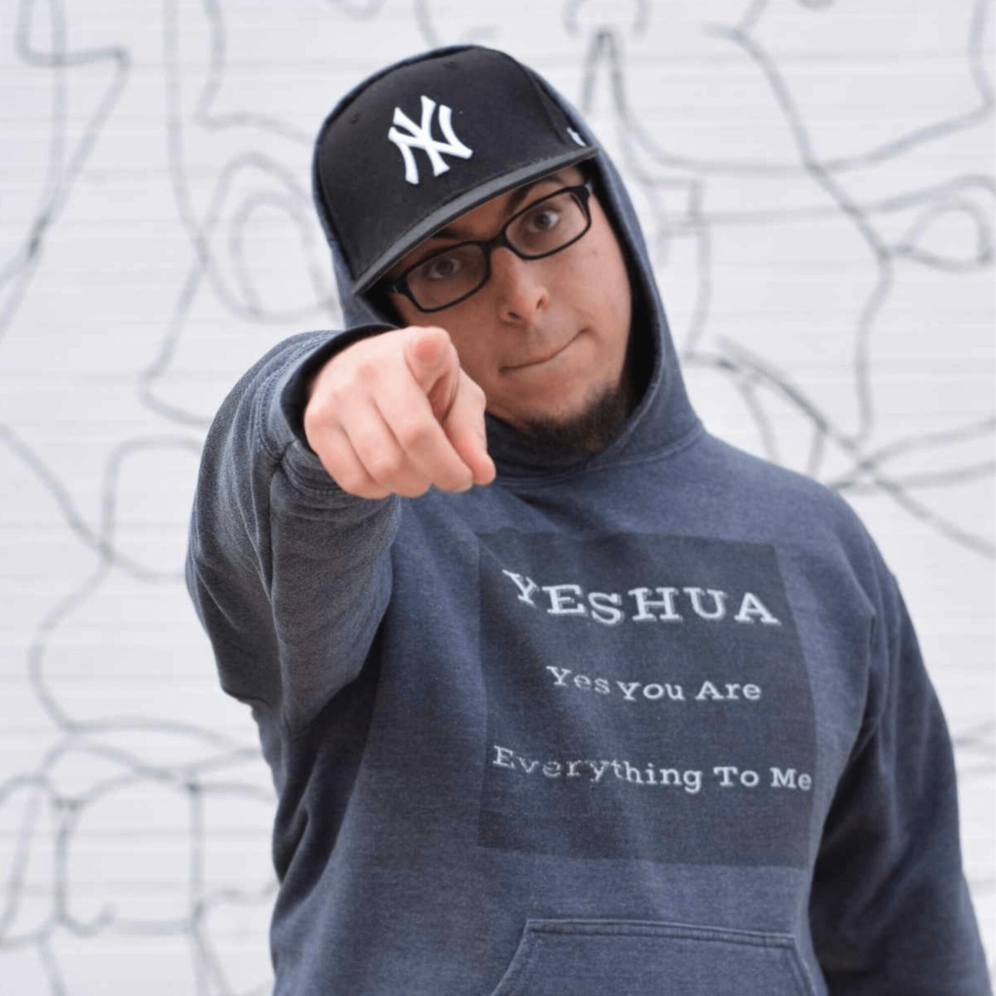 Person wearing glasses, a black New York Yankees cap, and a hoodie pointing at the camera, background with abstract lines.