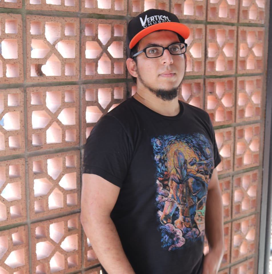 Young man with glasses and a beard wearing a black graphic t-shirt and a black and orange cap, standing against a brick wall.
