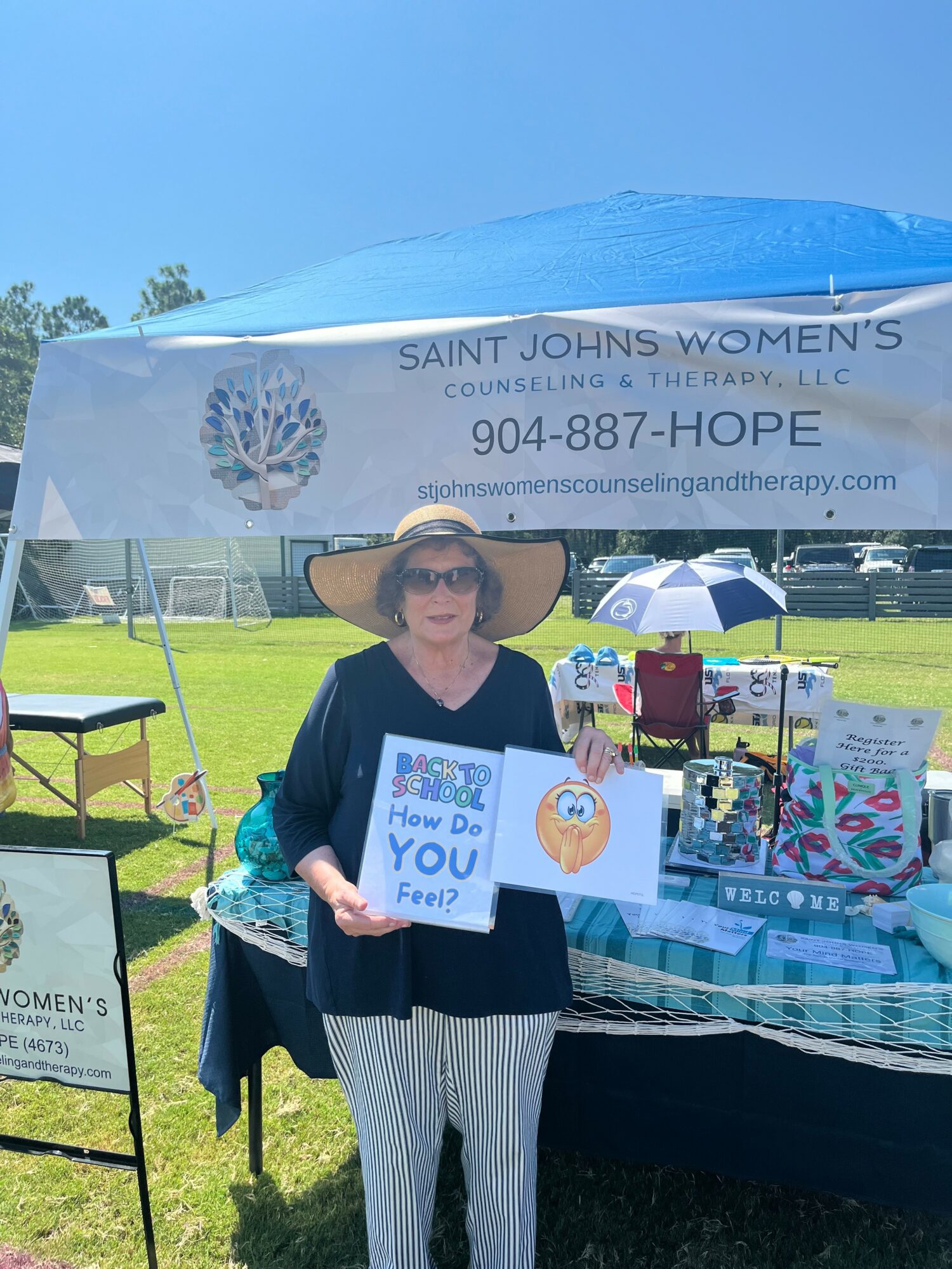 Woman standing outdoors near a table with informational materials, under a blue tent with a sign for Saint Johns Women's Counseling, holding a sign that says 