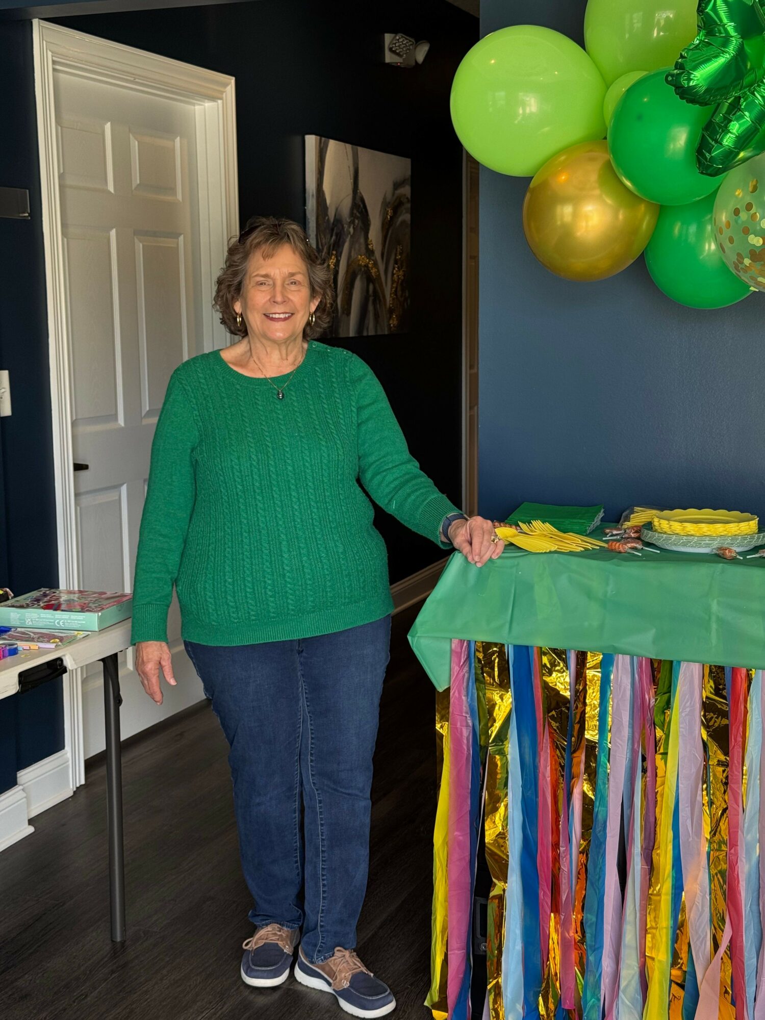 Woman standing indoors near a table with colorful decorations, holding a green gift bag, smiling, wearing a green sweater and jeans.