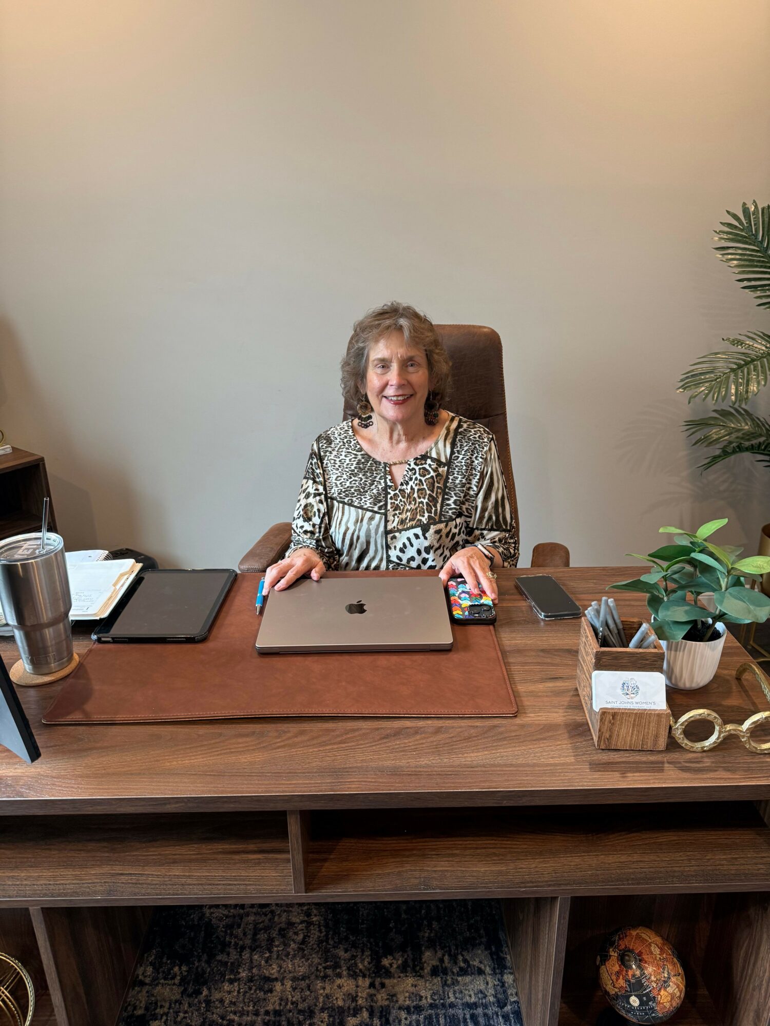 Woman sitting at a desk with a laptop, phone, and decorative items in a room with a bookshelf and plant.