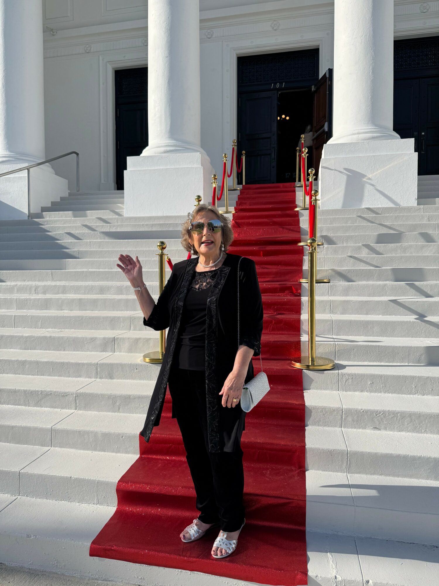 Older woman with sunglasses, black outfit, and white shoes standing on a red carpet outside a building with white columns.