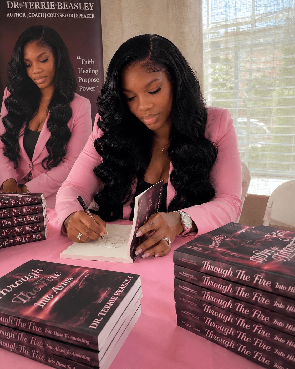 Woman with long black hair in pink blazer writing in a book, surrounded by stacked copies of her book, in a bright room.