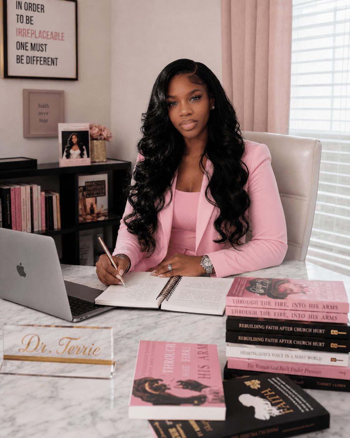 Woman with long black hair in pink blazer sitting at desk with books, laptop, and notepad in a bright room.