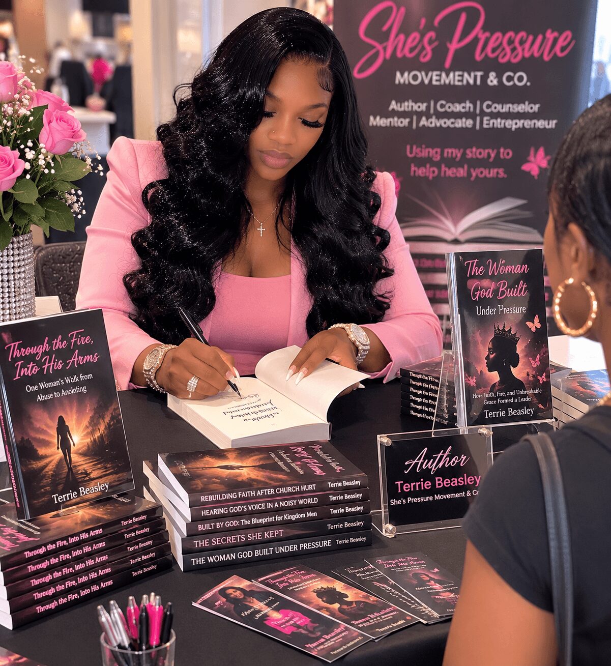 Woman with long black hair in pink top signing books at a table, promotional materials and books displayed, pink background.