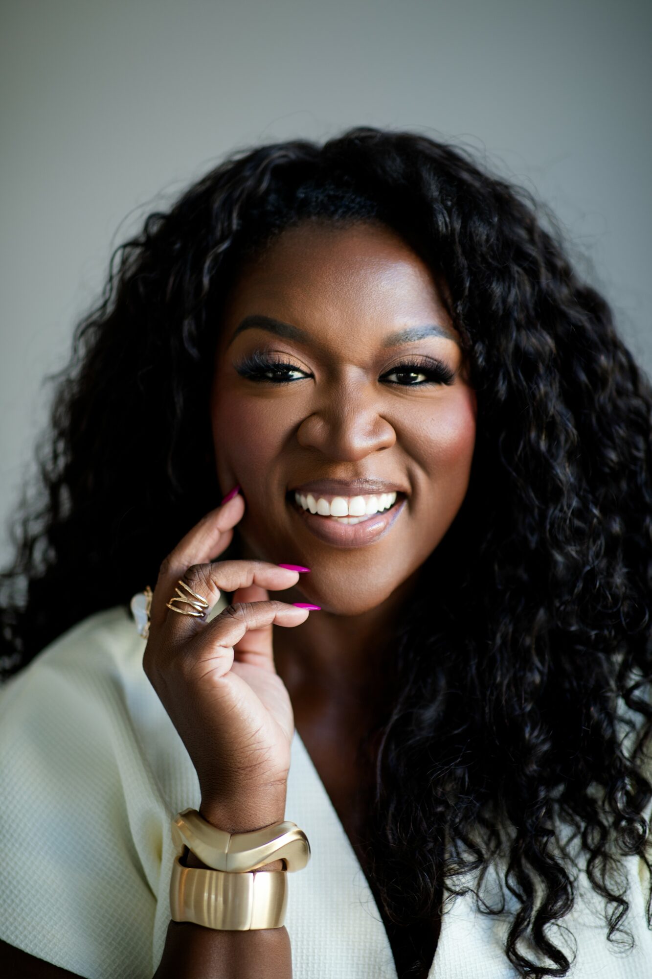 Woman with curly hair smiling, wearing a white top and gold accessories, touching her face.