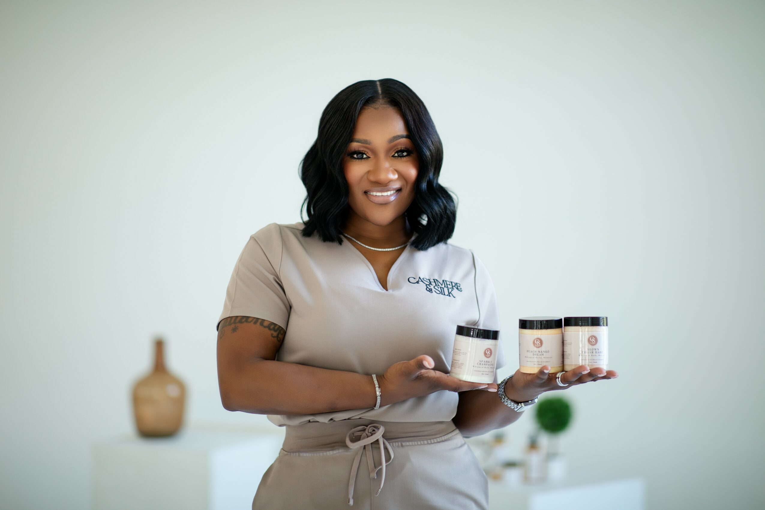 Woman with dark hair smiling, holding three jars, standing in a light-colored room with a blurred background.