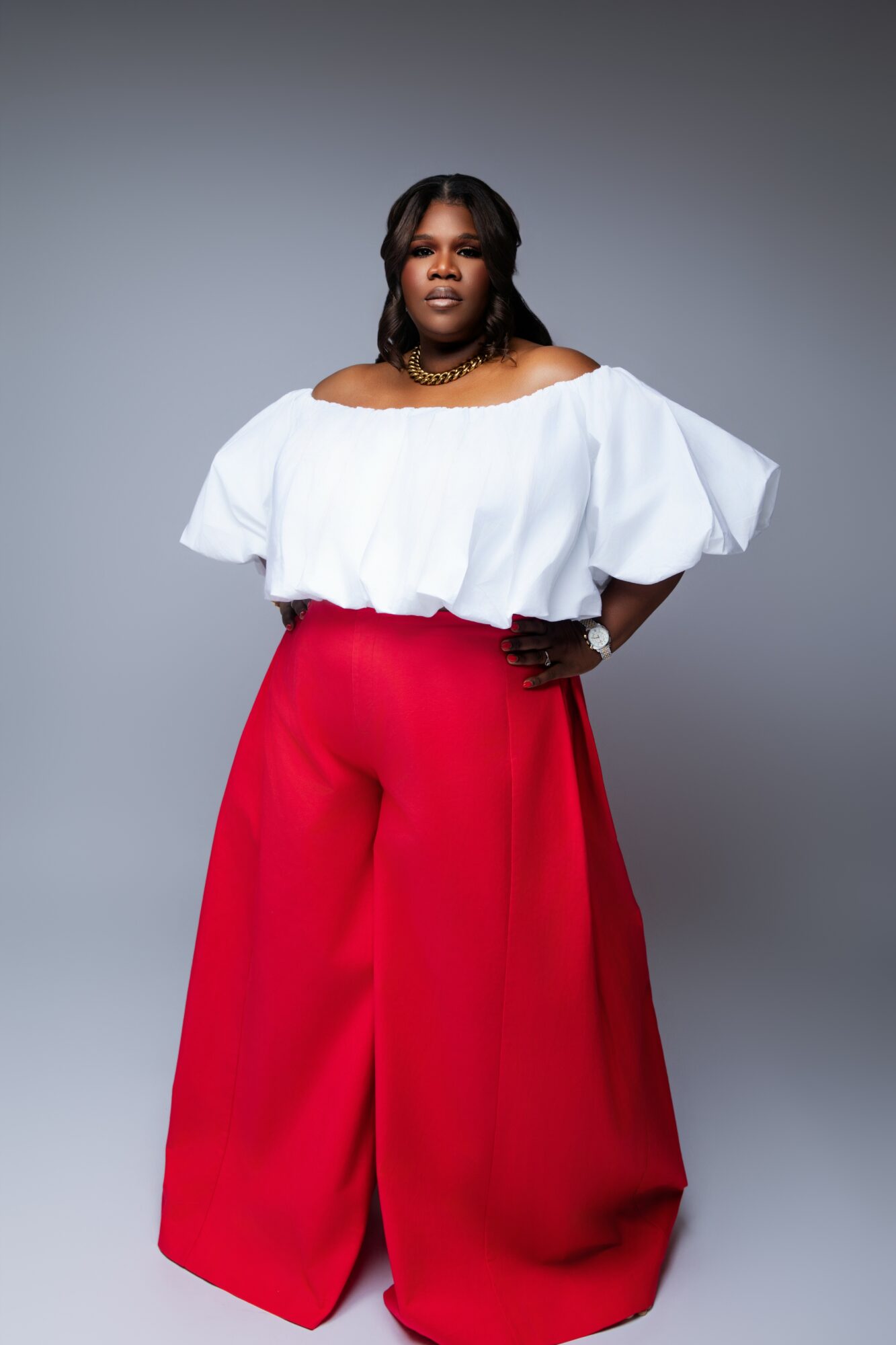 Woman in white top and red wide-leg pants standing against gray background, looking at camera with hand on hip.