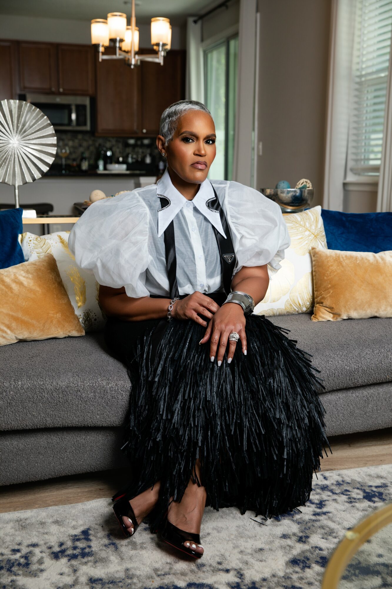 Woman sitting on a sofa with pillows in a living room, wearing a white puff-sleeve top and black feather skirt.