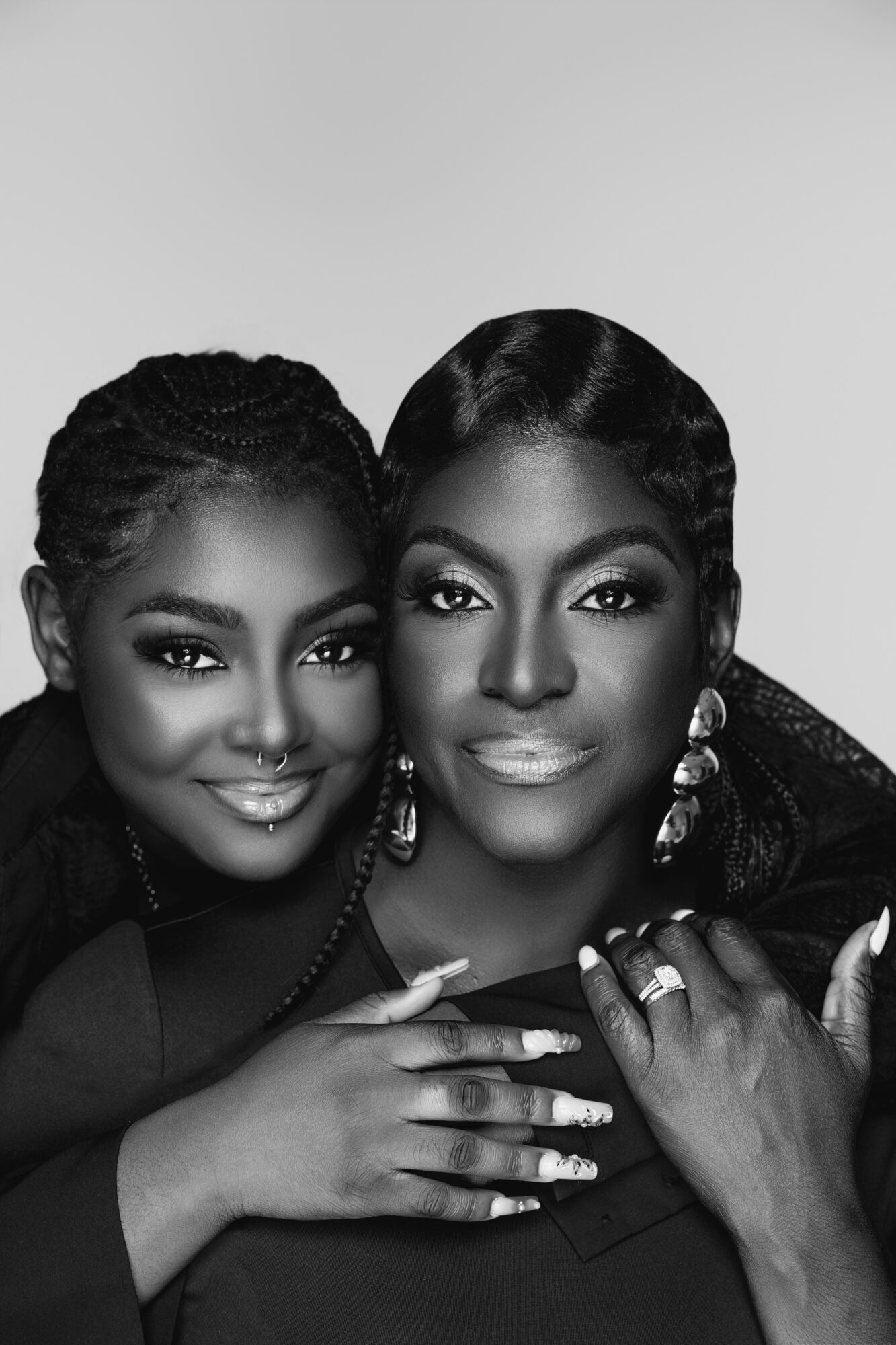 Two women with styled hair and jewelry, smiling closely together, in black clothing, against a plain background.