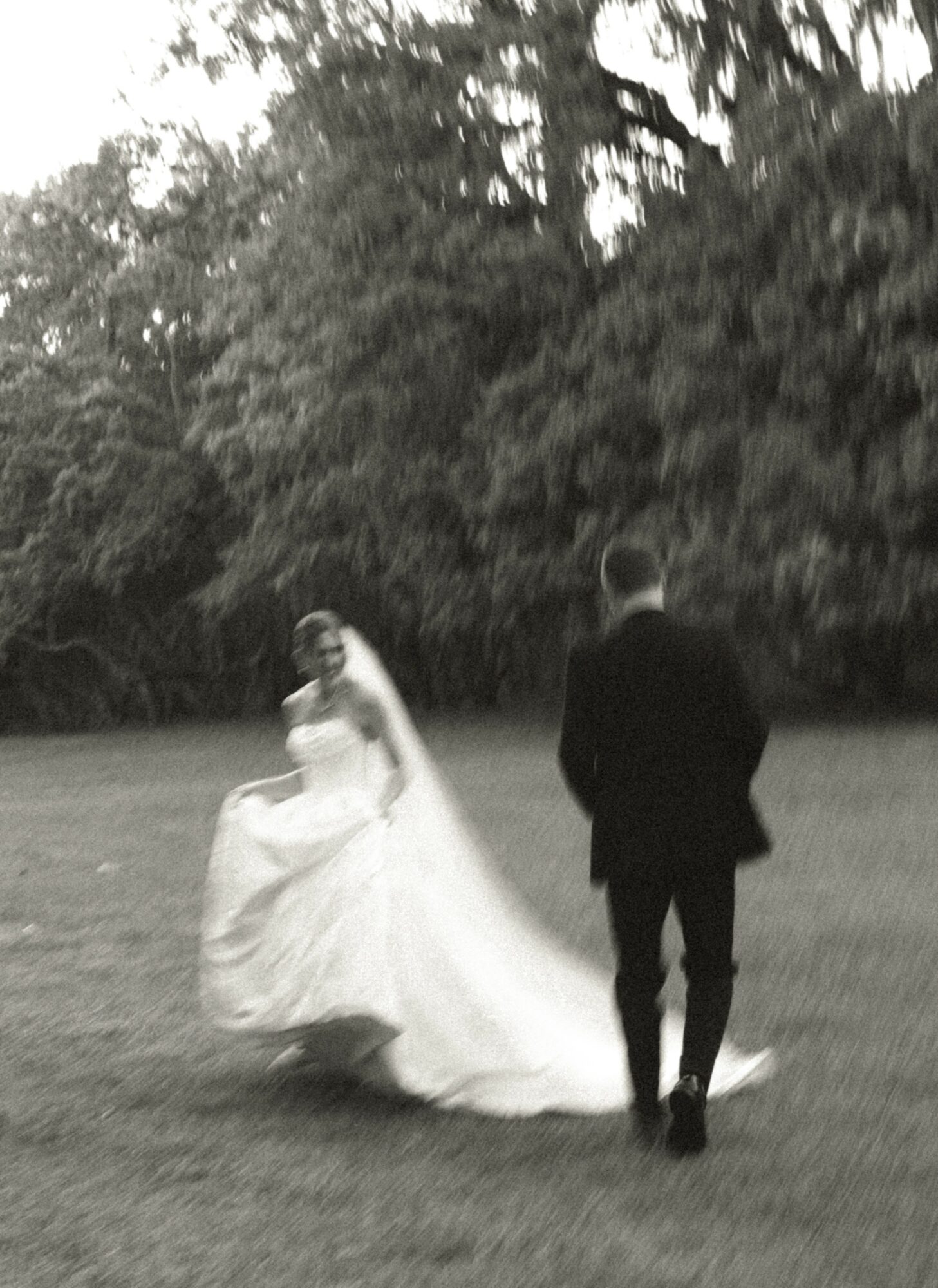 A bride in a wedding dress and a groom in a suit stand on grass with trees in the background.