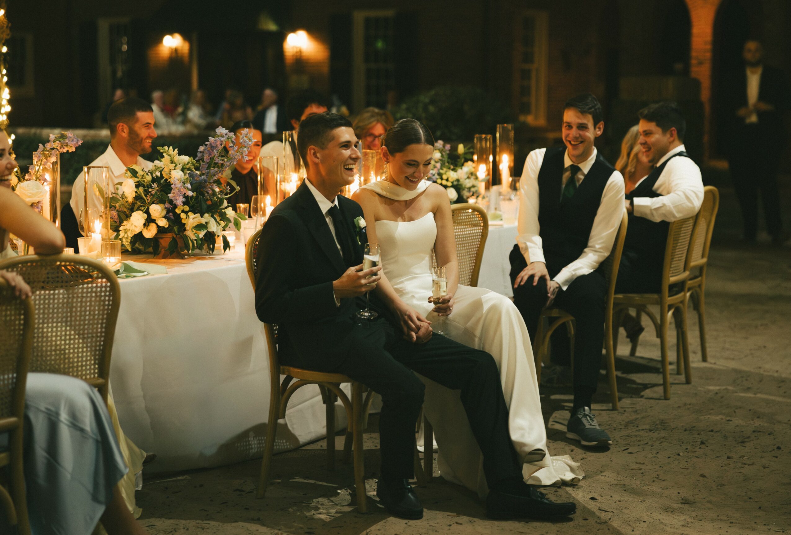 People seated at a wedding reception table, with floral arrangements and candles, in a dimly lit venue.