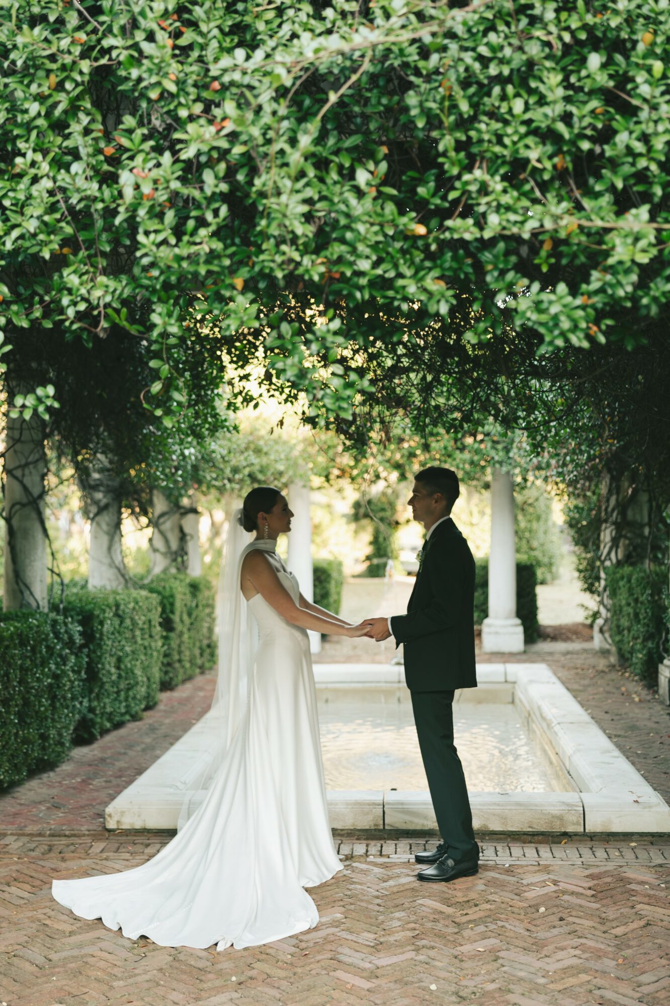A bride and groom holding hands in a garden with trees and a fountain, dressed in wedding attire.