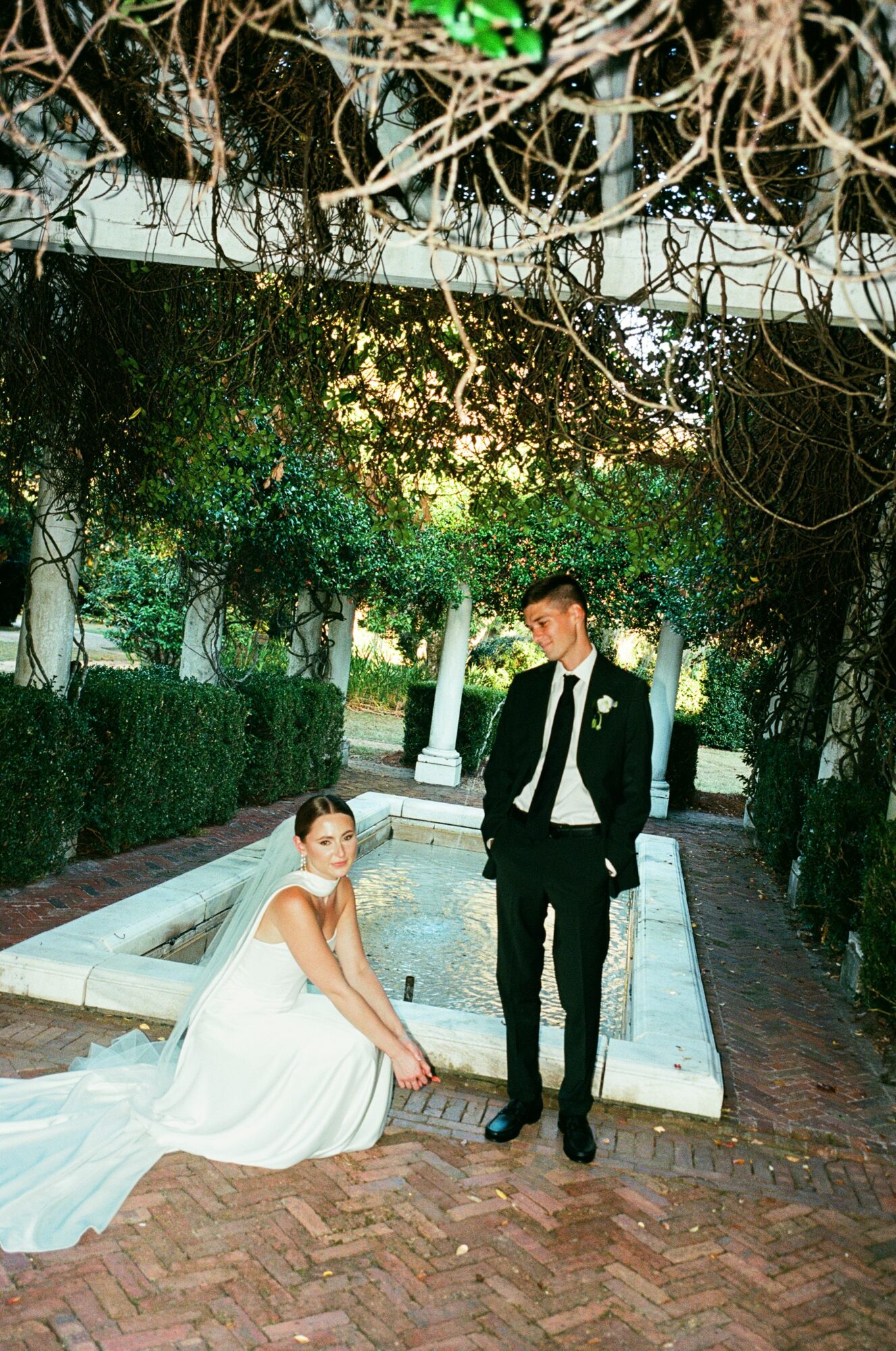Bride sitting on ground in white dress, groom standing beside her in suit, outdoors with trees and fountain background.