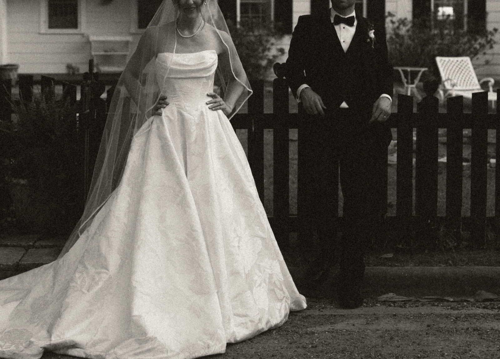 Bride in a wedding dress and veil standing next to a groom in a tuxedo outdoors, with a fence and house in background.