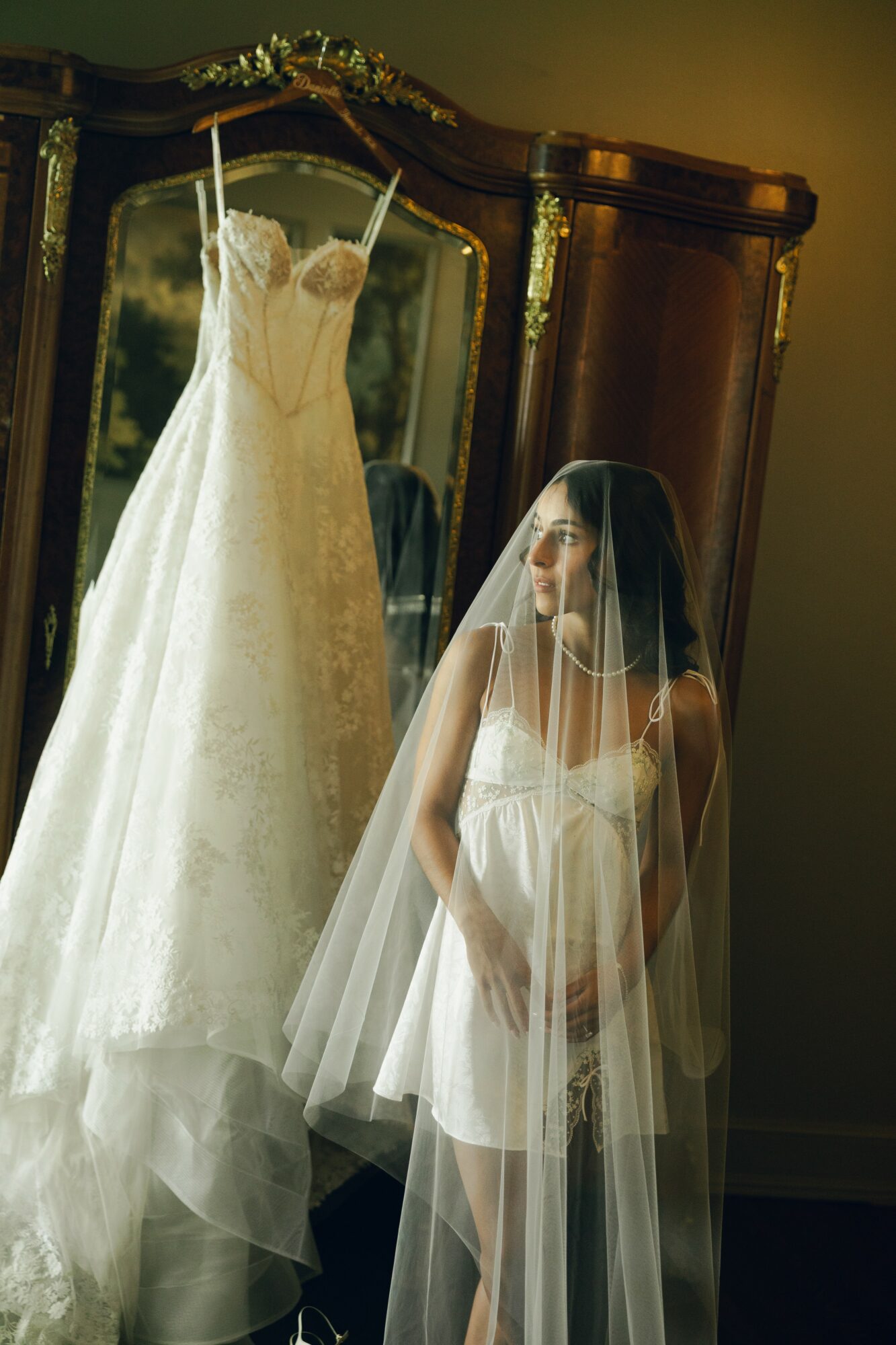 Woman in wedding dress with veil looking at a wedding gown in a mirror, standing indoors. The gown is on a hanger.