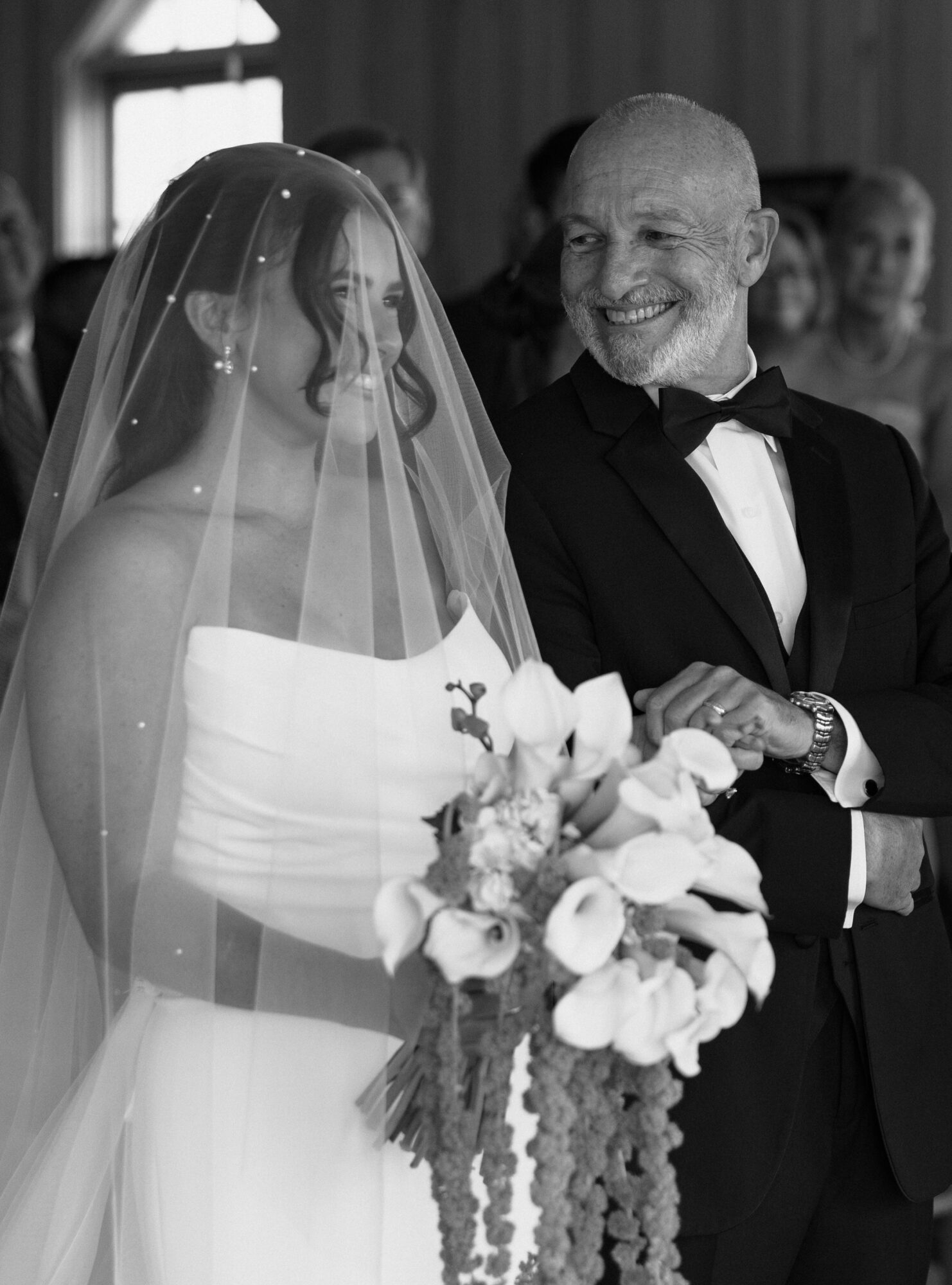 A bride and an older man, possibly her father, smiling at each other during a wedding ceremony, with the bride holding a bouquet.