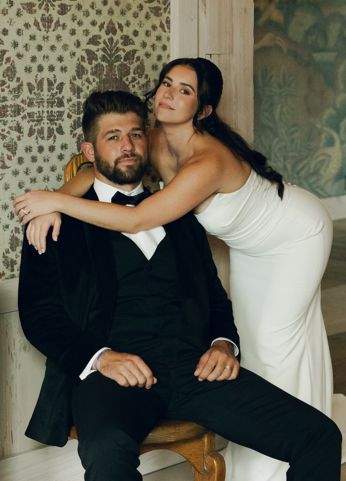 A woman in a white dress leans over a seated man in a black tuxedo, both in an elegant setting.