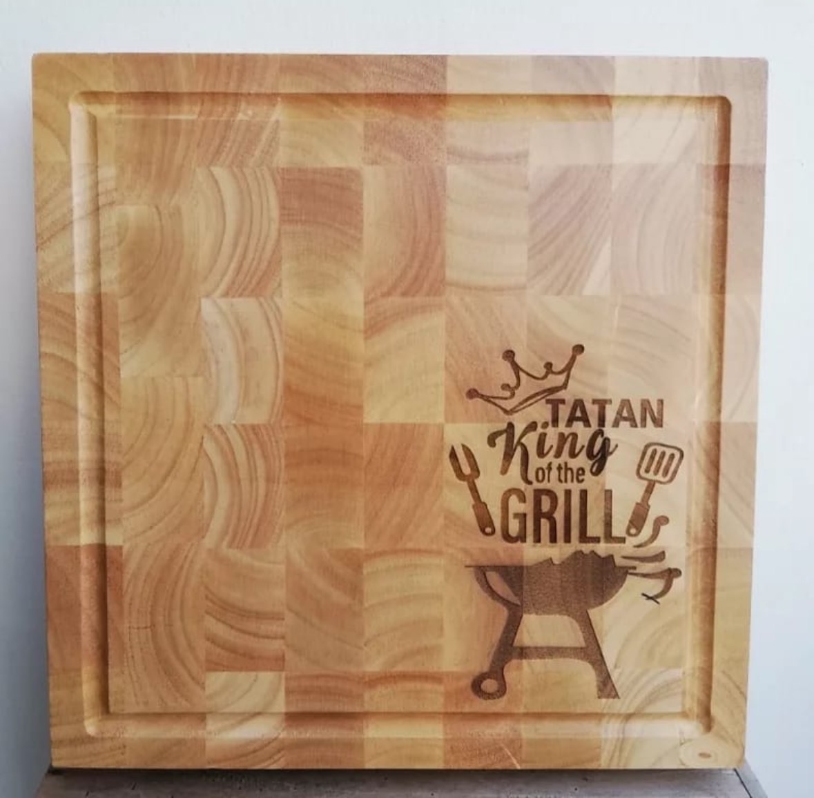Wooden cutting board with engraved text and grill illustration, reading 'Tatan King at the Grill' with a crown, fork, and spatula.