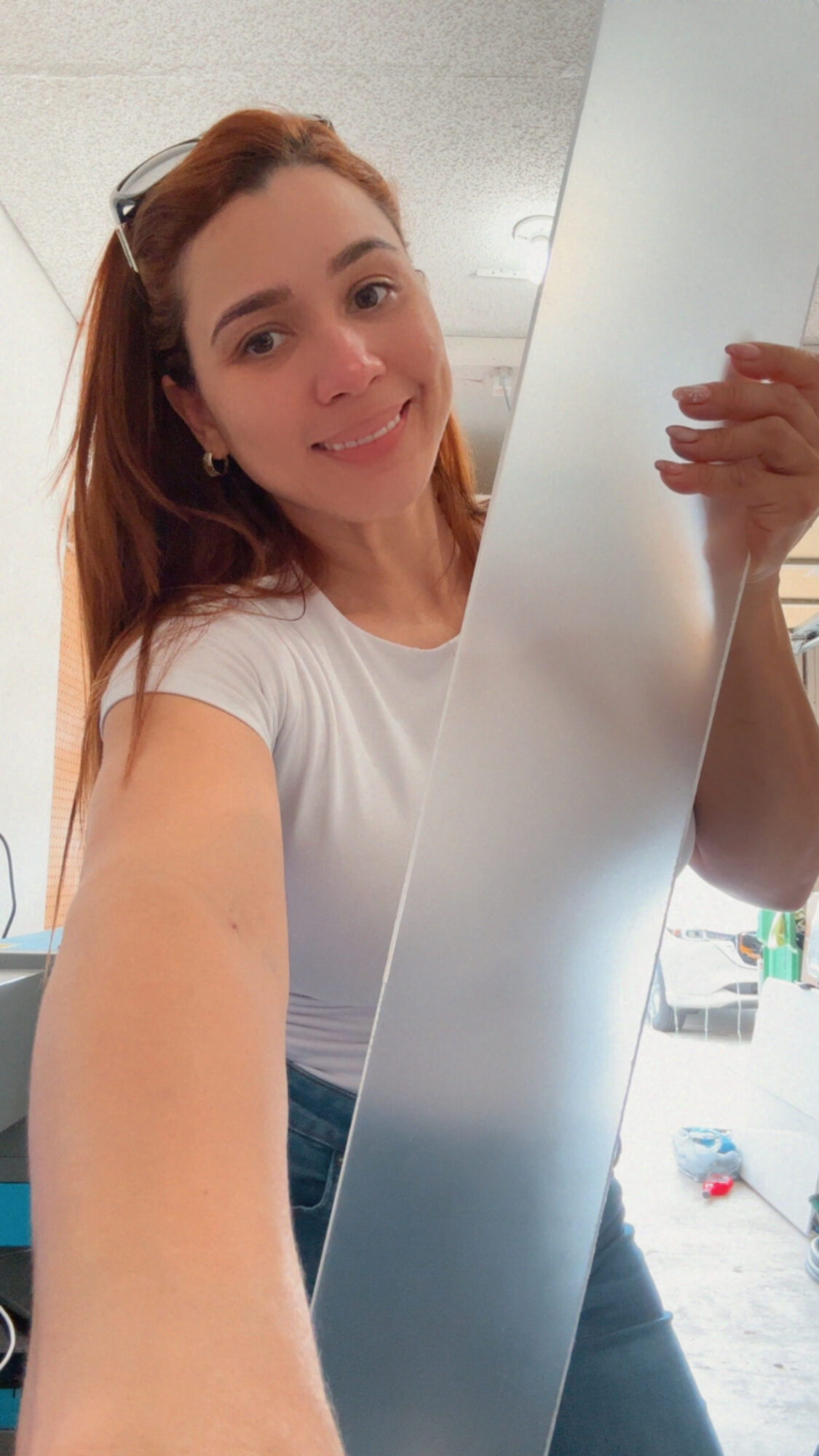Young woman with long hair taking a selfie in mirror, smiling, wearing a white shirt, in a bright room.