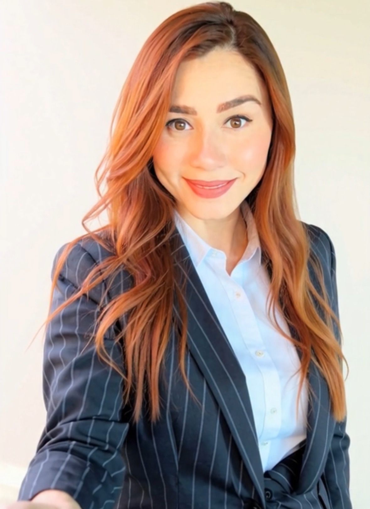 Young woman with long, wavy reddish-brown hair, smiling, wearing a pinstripe blazer and light blue shirt, against a light background.
