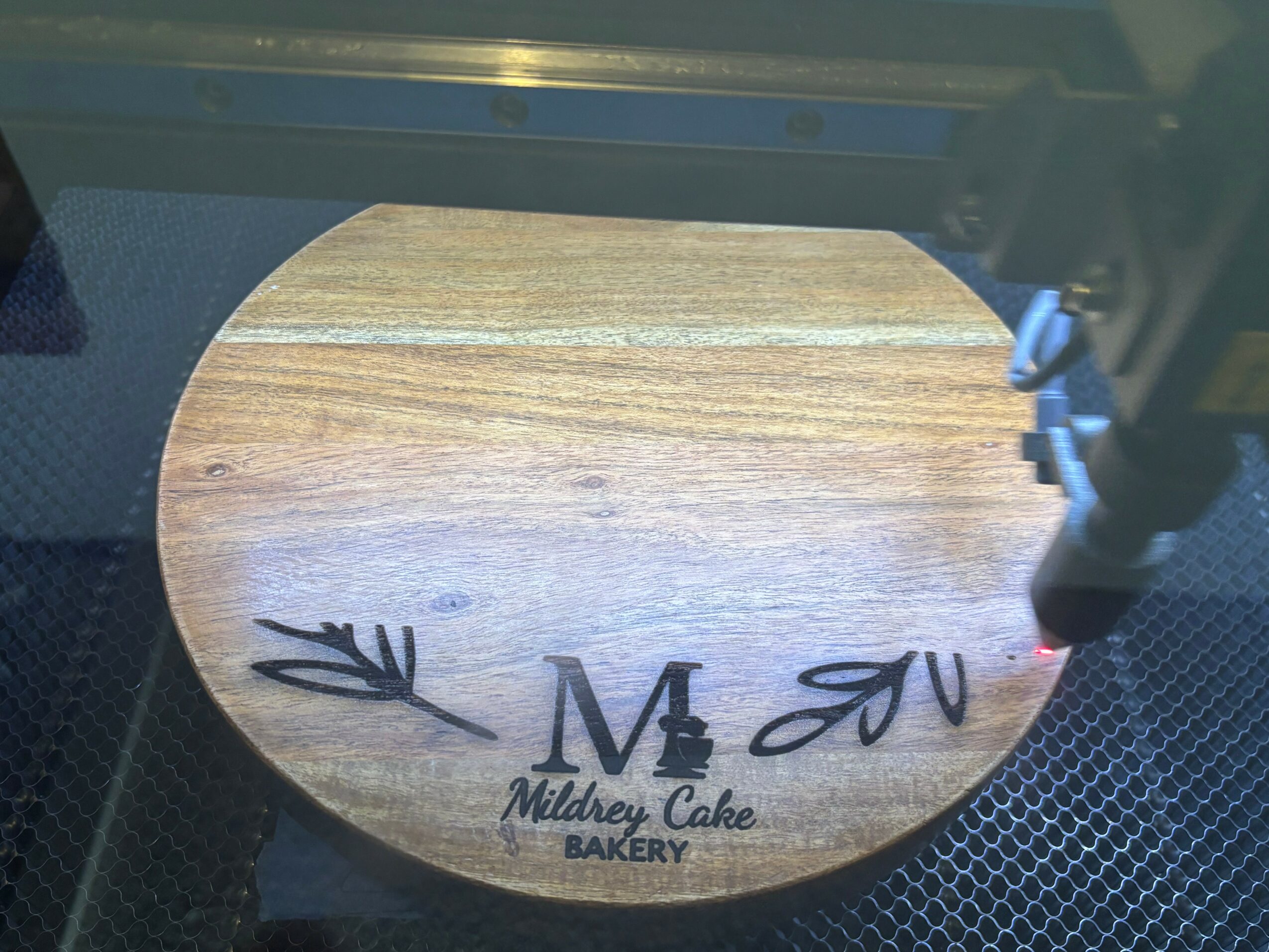 Round wooden cake board with initials and decorative leaves, with bakery logo at bottom, viewed from the side.