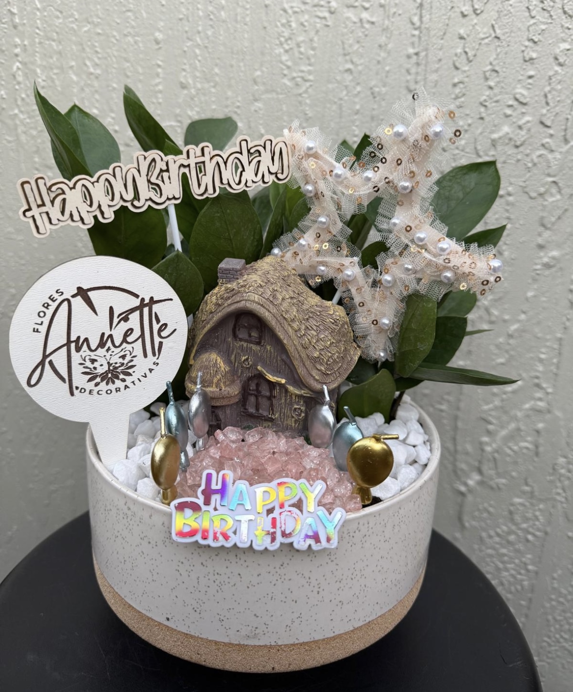 Decorative birthday arrangement with a small house, greenery, and birthday signs in a round container.