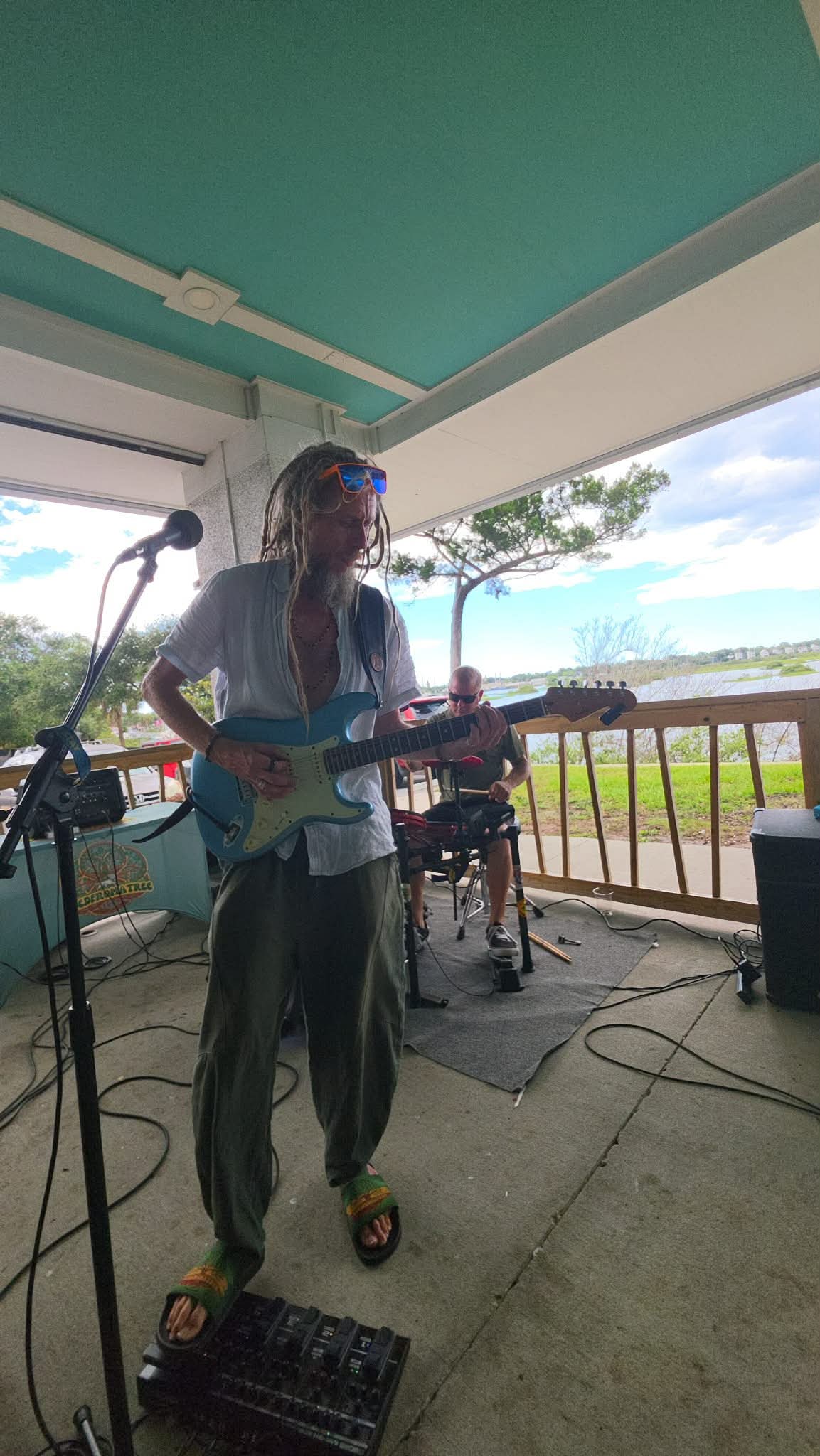 Person playing electric guitar on porch with ocean view, trees, and sky in background.