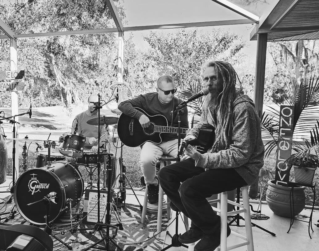 Two musicians perform outdoors with guitars, drums, and microphones under a canopy, surrounded by trees and plants.