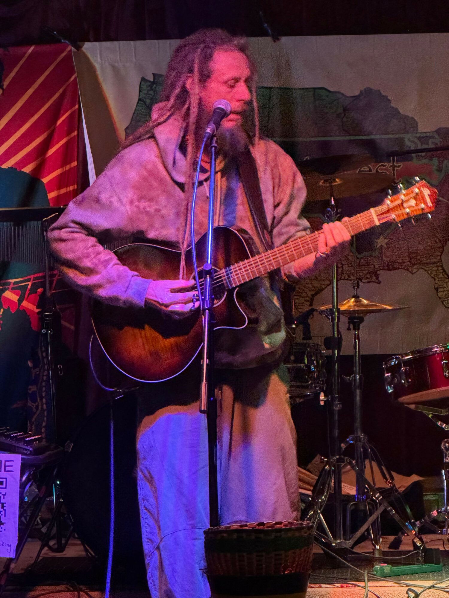 Person singing and playing guitar on stage with microphone, wearing a fur coat and light-colored pants, in a performance setting.
