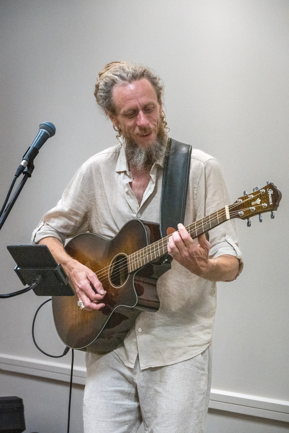 Man with long hair and beard playing an acoustic guitar, standing near a microphone, in a plain room.