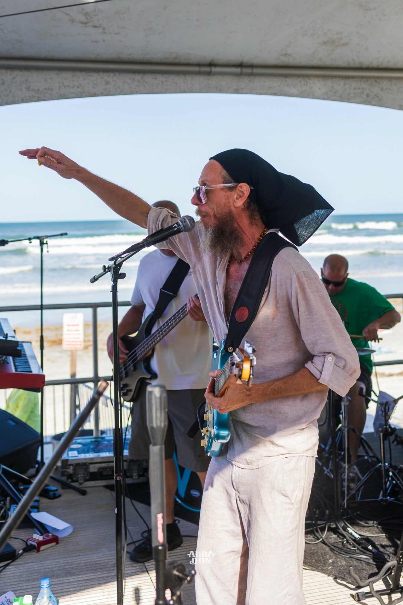 A man with a beard, wearing sunglasses and a head covering, plays guitar and sings into a microphone on a beachside stage.