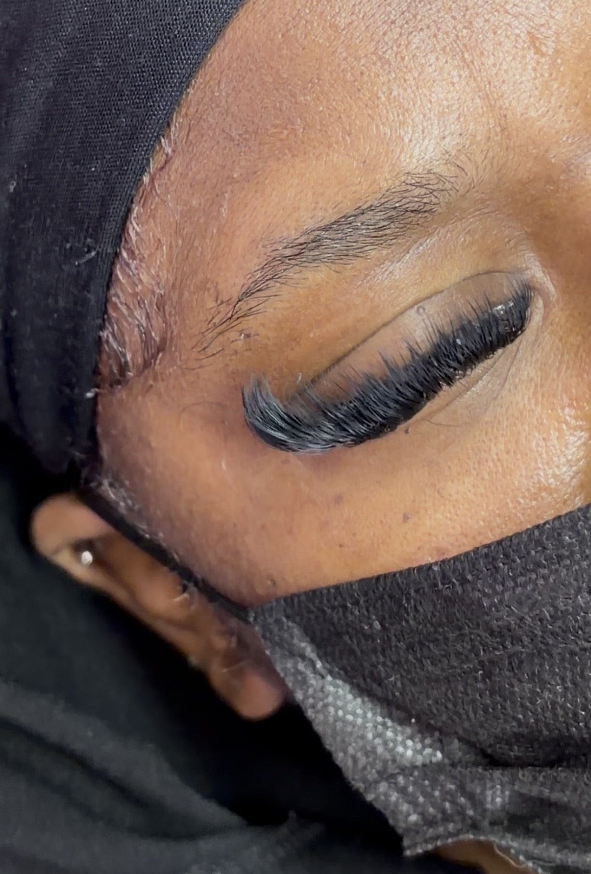 Close-up of a person's eye with long eyelashes, partially covered by a black face mask and head covering.
