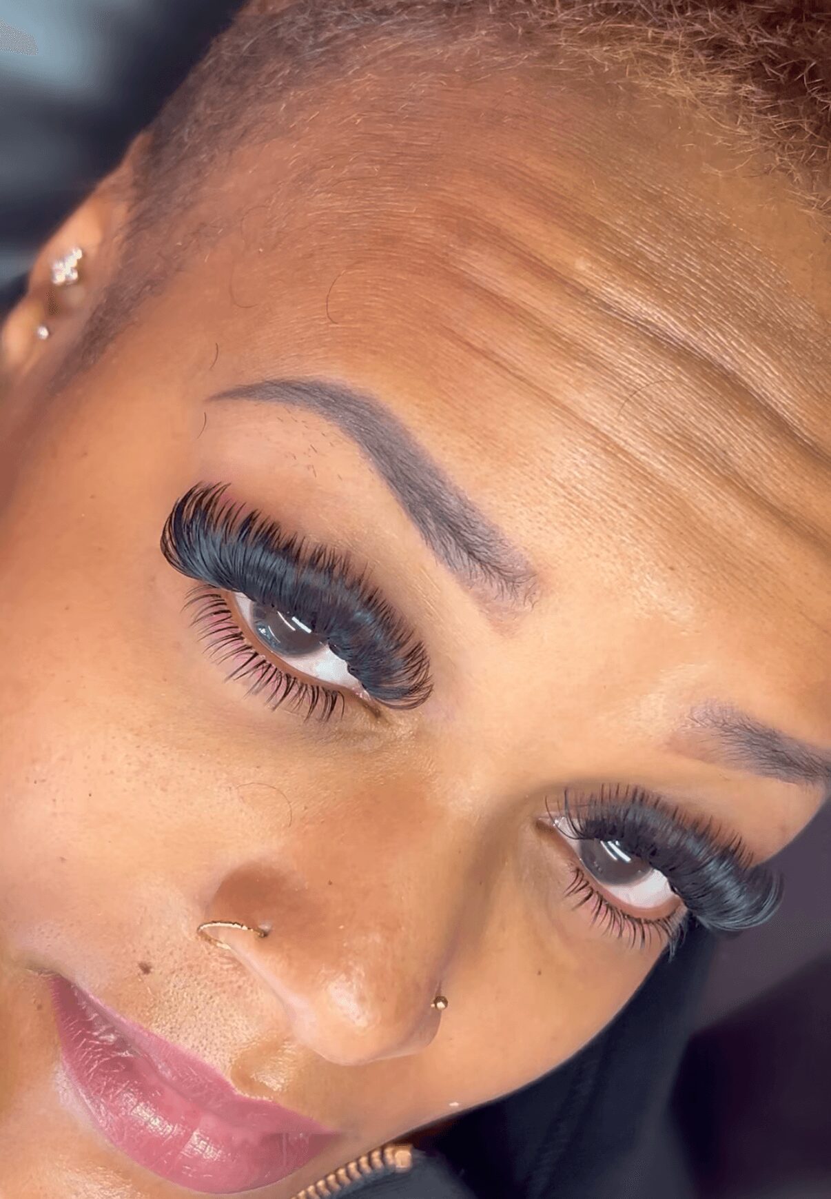Close-up of a woman's face with makeup, including long eyelashes, eyebrows, and a nose piercing.