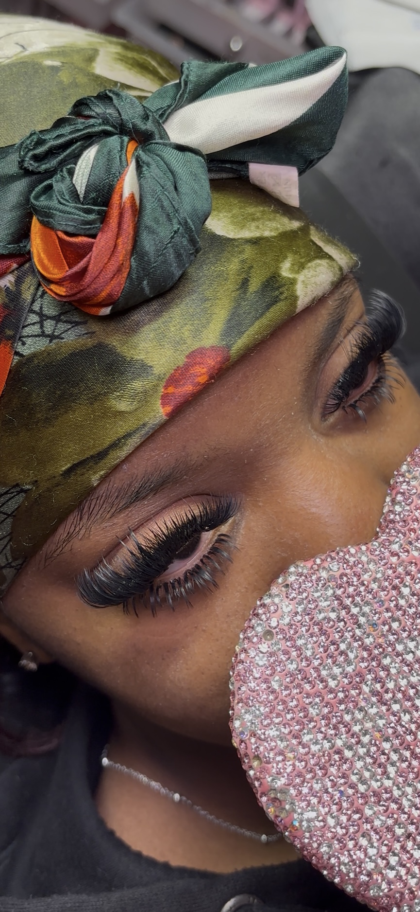 Close-up of a woman's eyes with makeup, wearing a colorful headscarf, and holding a pink glittery object near her face.