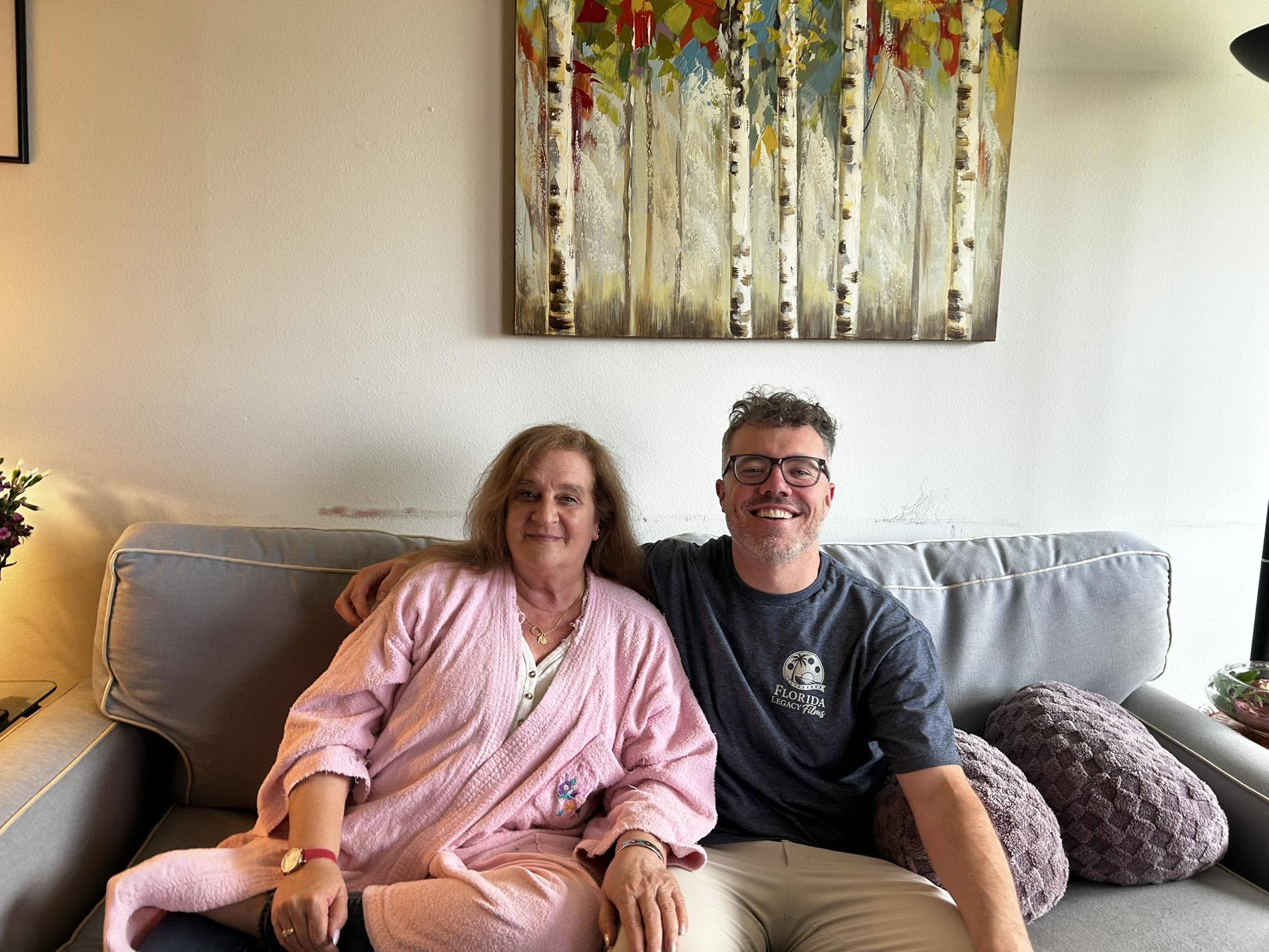 Two people sitting on a sofa, smiling, with a colorful painting above them, in a cozy room.