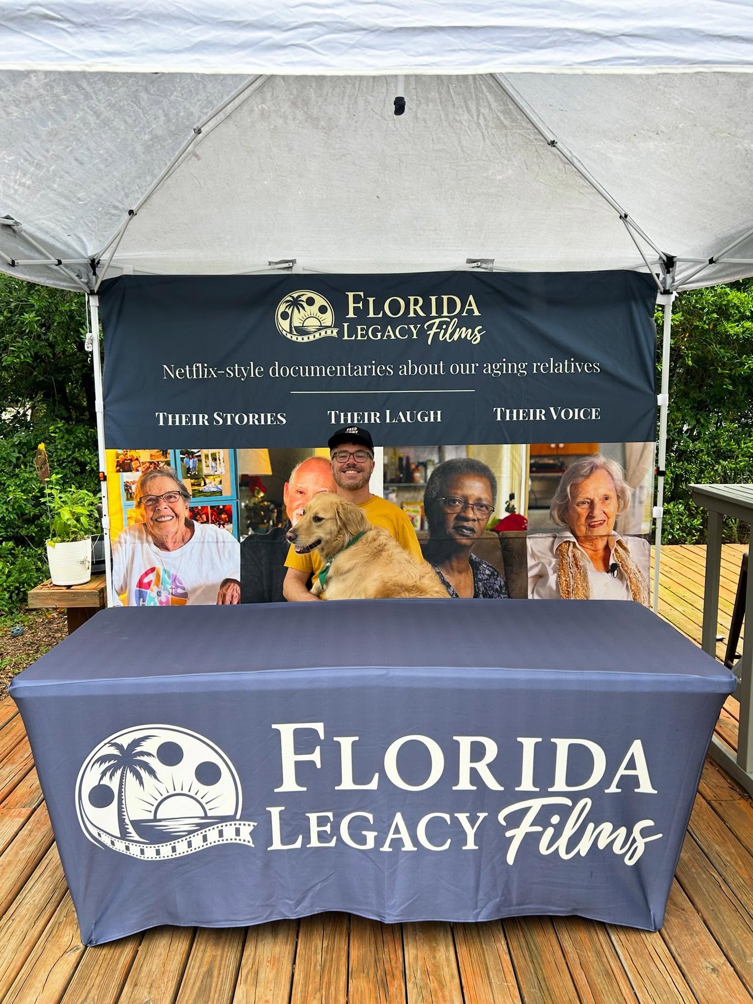 Table with Florida Legacy Films logo, under a canopy, with four people and a dog behind it, outdoor setting.