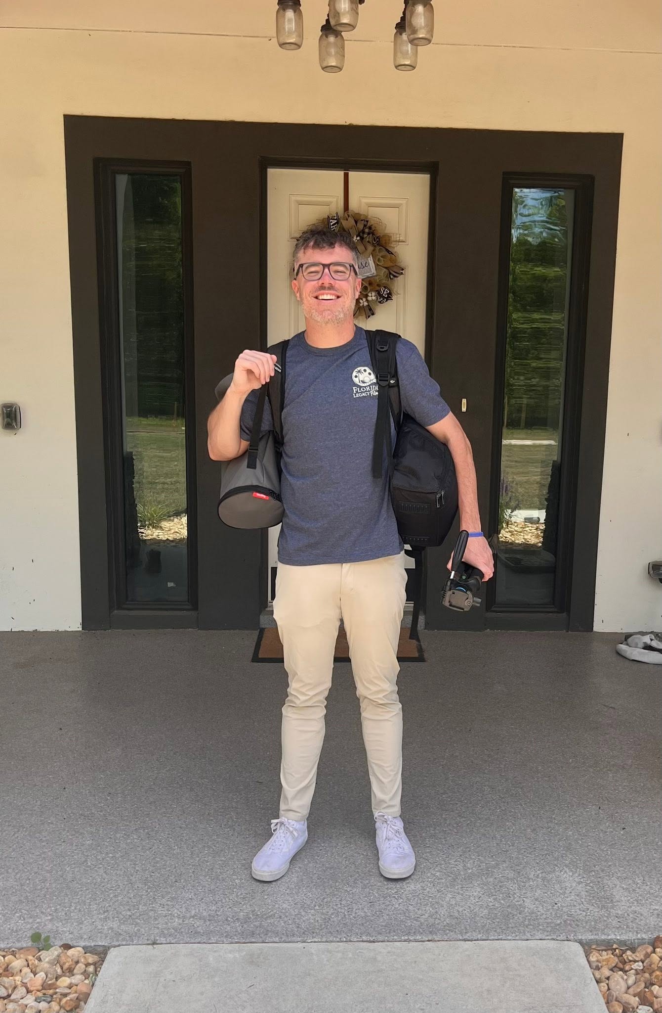 Person standing outside in front of a building, smiling, wearing glasses, a backpack, and casual clothes, holding a camera.