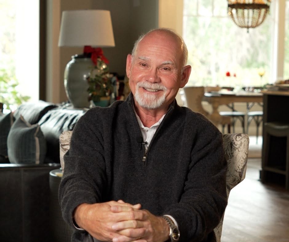 Older man with a beard smiling, sitting indoors with a living room background, including a lamp and window.