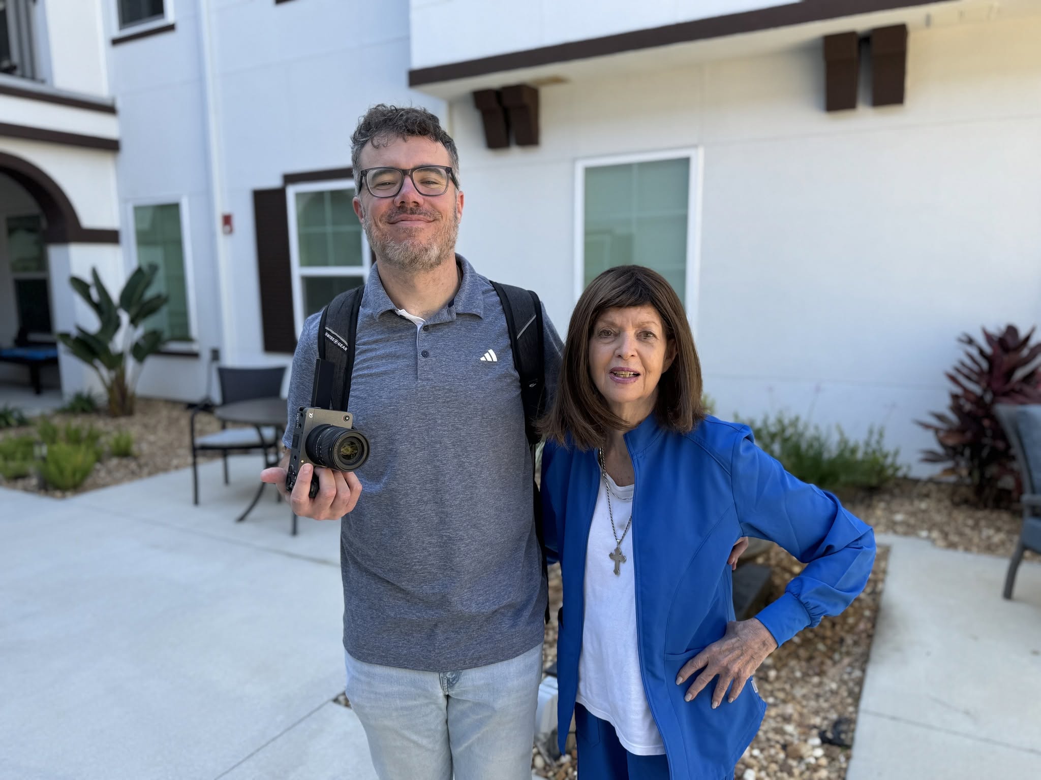 A man and woman stand outdoors in front of a white building, smiling, with the man holding a camera, both wearing casual clothing.