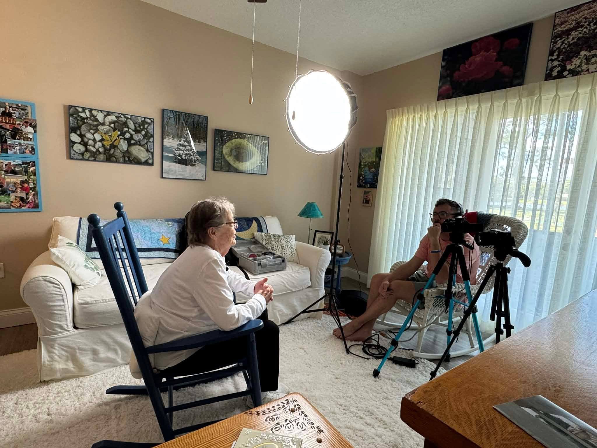 Two women sit in a living room with a white sofa, wall art, and a large window with curtains. One woman is in a white shirt, the other in casual attire, with a camera on a tripod nearby.