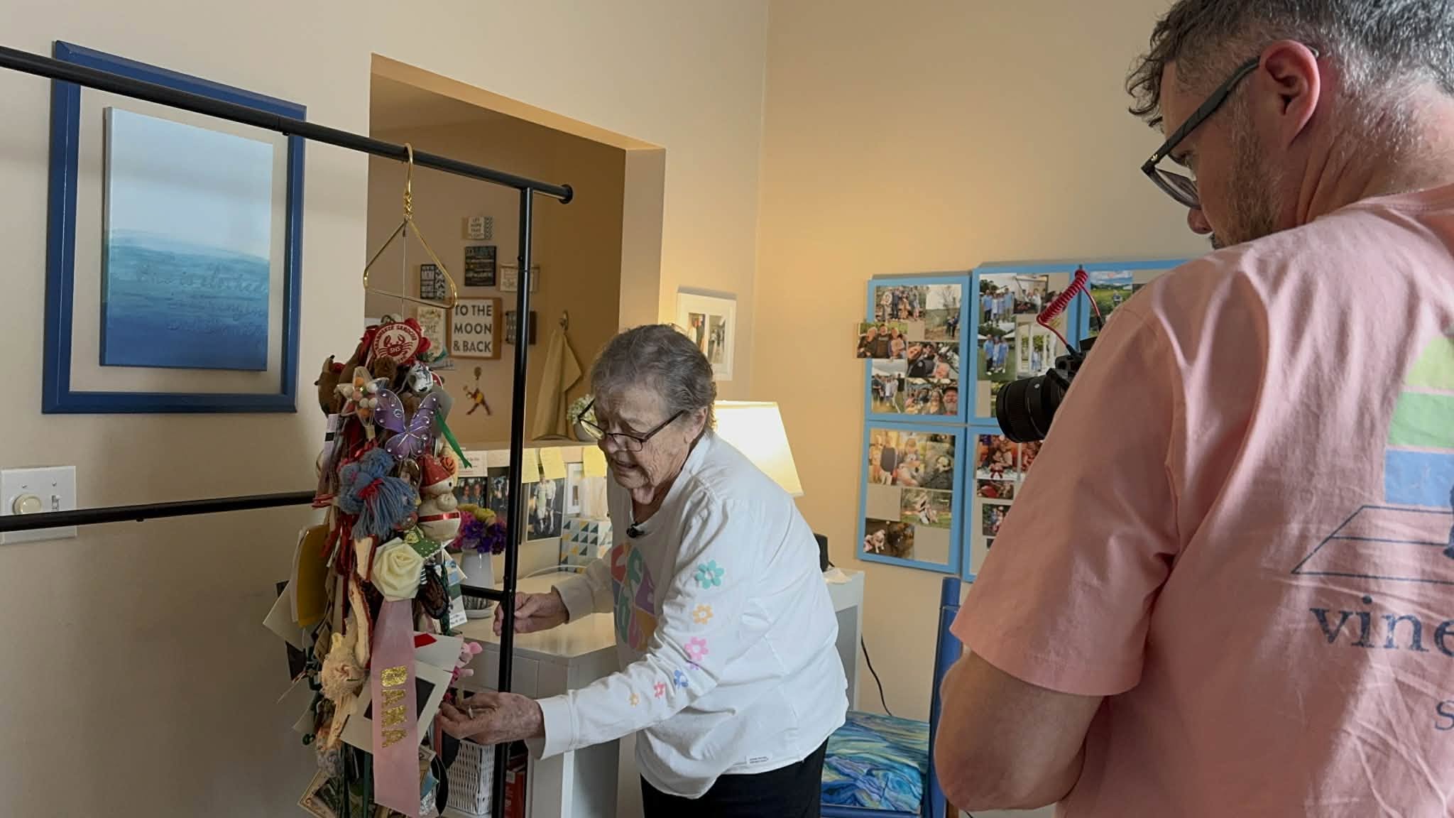 Two people in a room with artwork and photos, one woman arranging flowers, one man observing, both wearing glasses.
