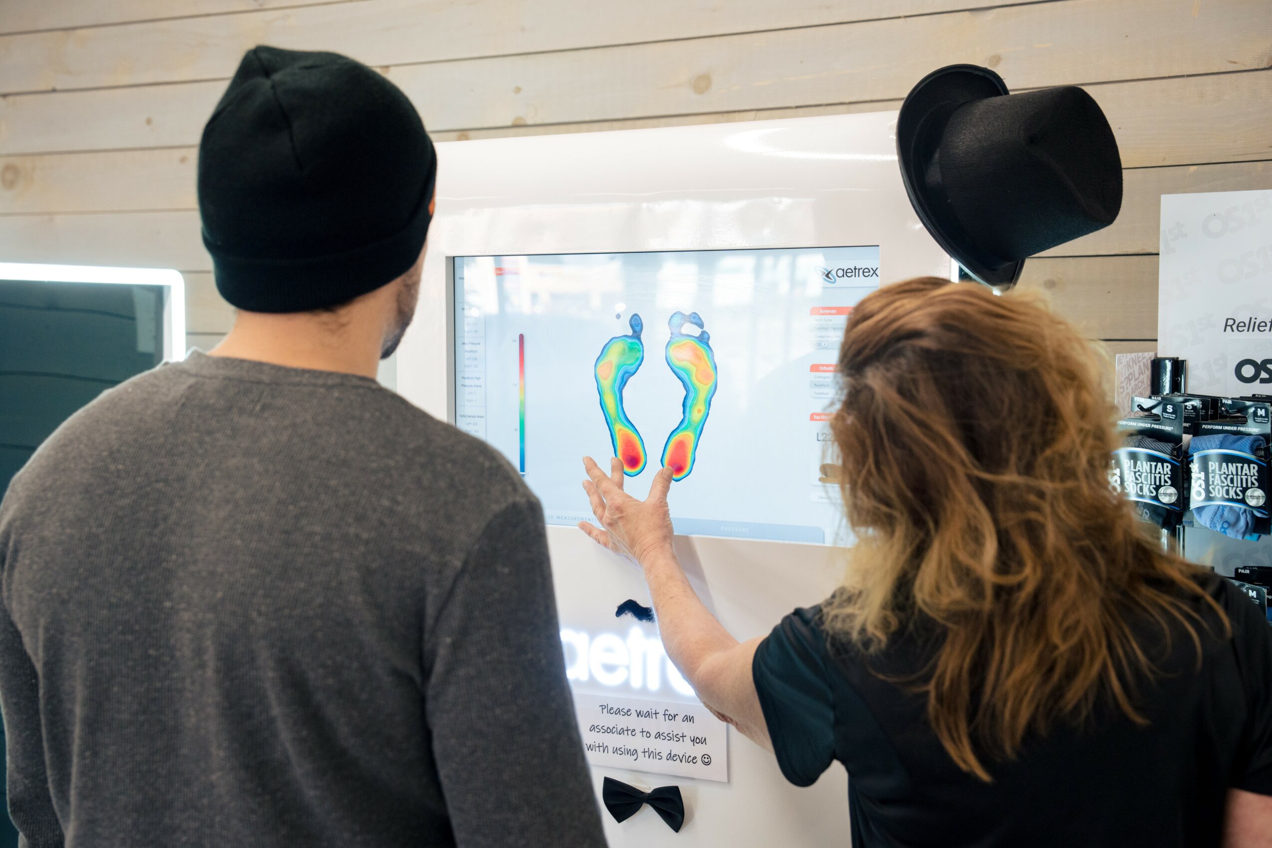 Two people, one with a black hat, look at a screen displaying colorful foot scans in a room with wooden walls.