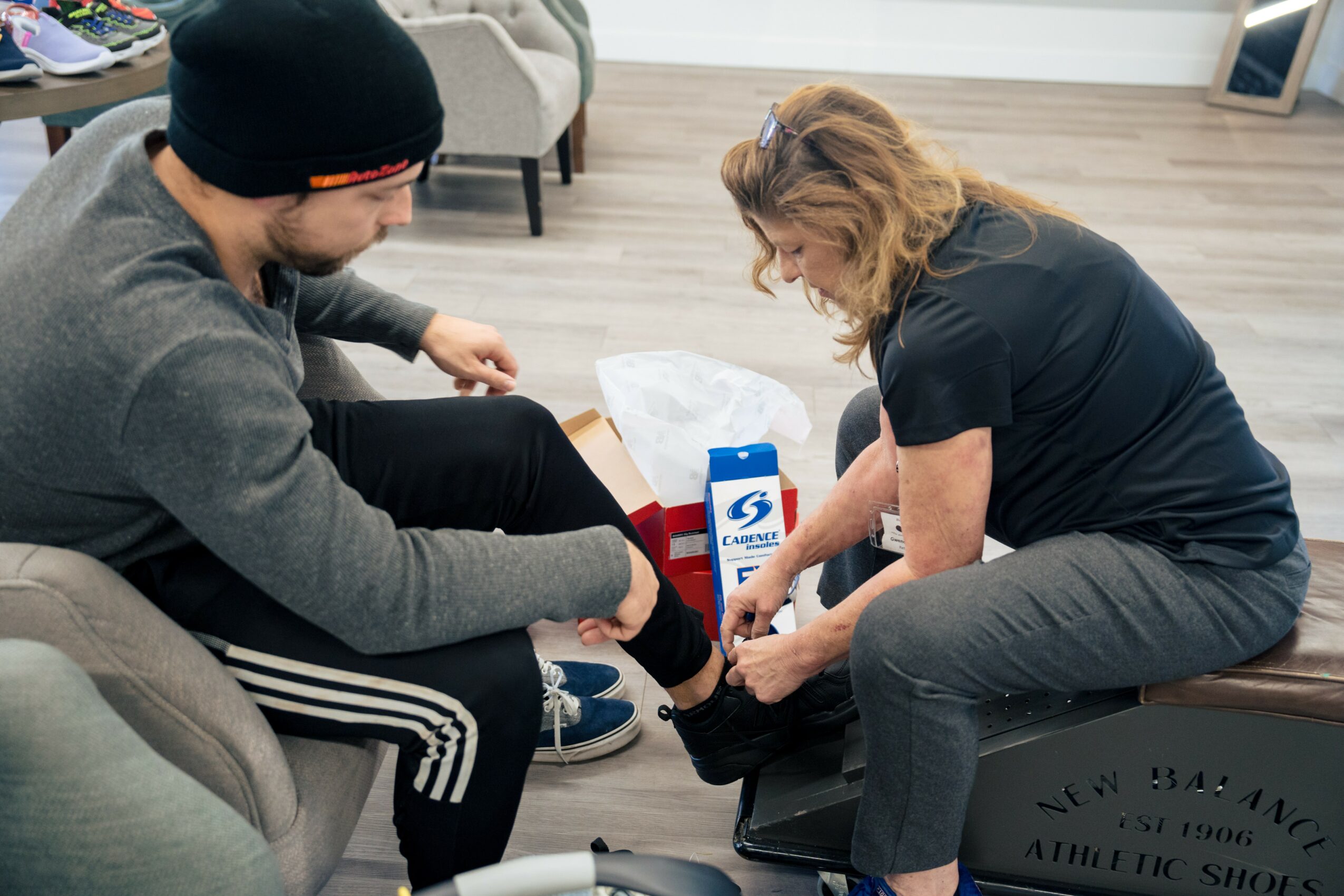 Two people sitting on a couch, one helping the other with a shoe, in a living room setting.