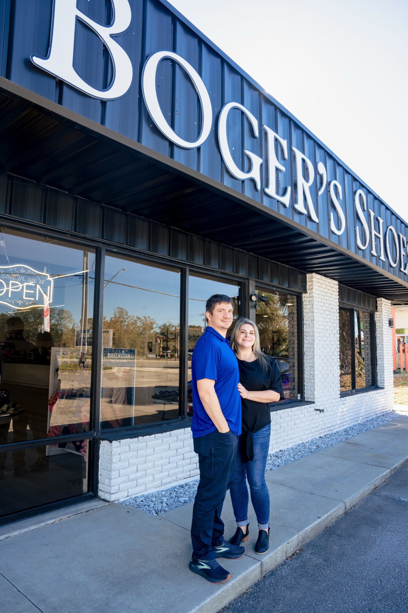 Two people stand outside a store with a large sign reading 'Boger's Shop'. They are smiling and posing for the photo.