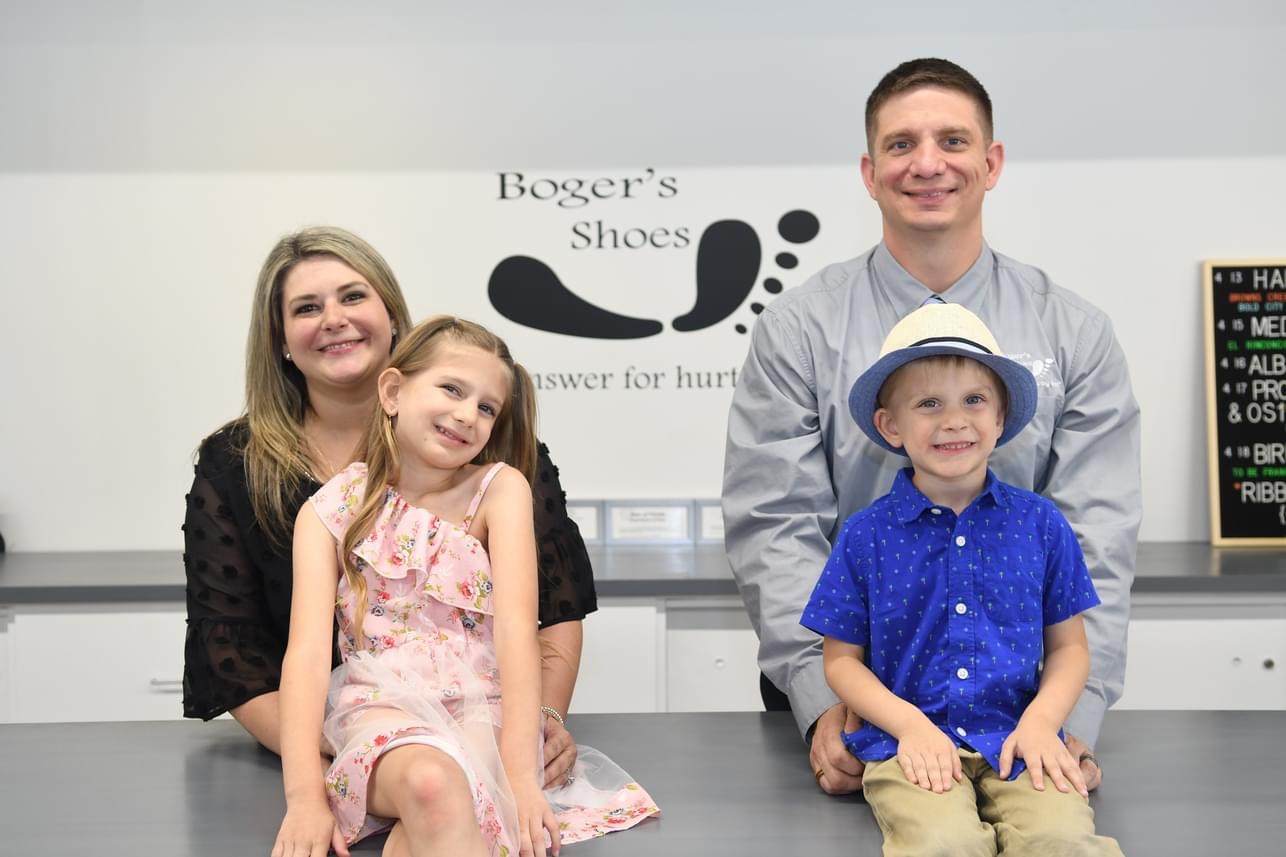Family of four smiling, two children and two adults, standing behind a counter in a shoe store, with a sign reading 'Boger's Shoes'.