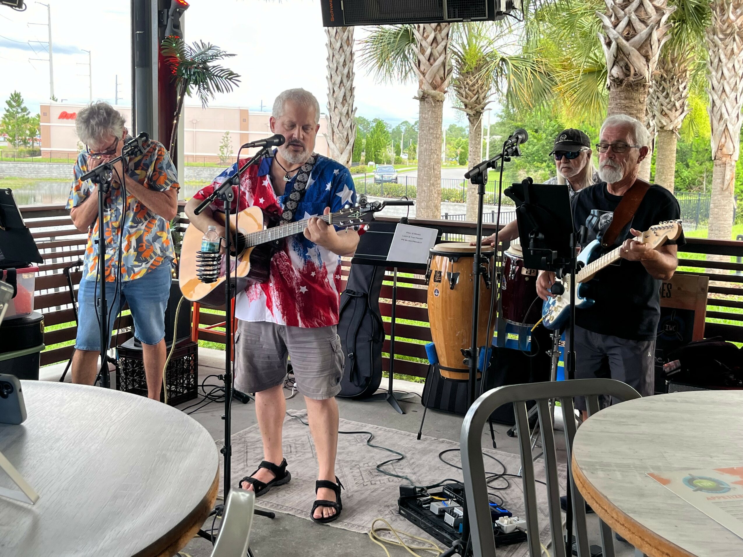 Four musicians perform outdoors with guitars, percussion, and microphones, surrounded by palm trees and seating areas.